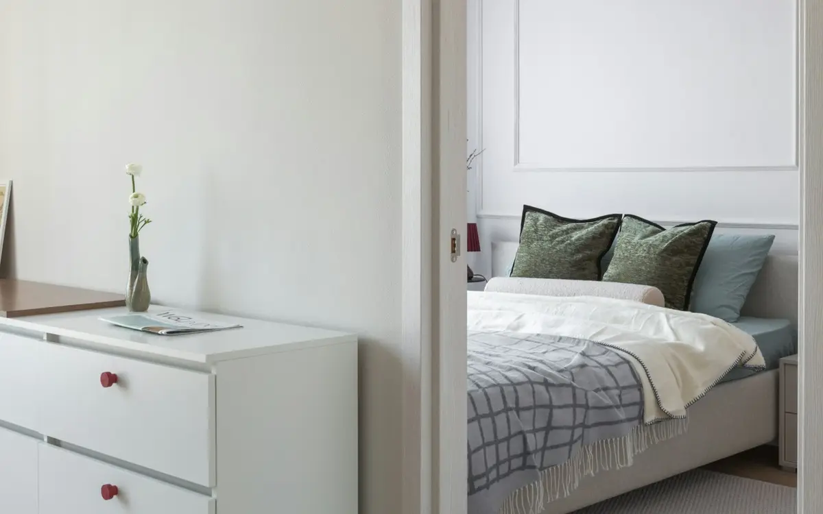 A bedroom seen through an open door, with a chest of drawers in the foreground and a bed on a solid upholstered base