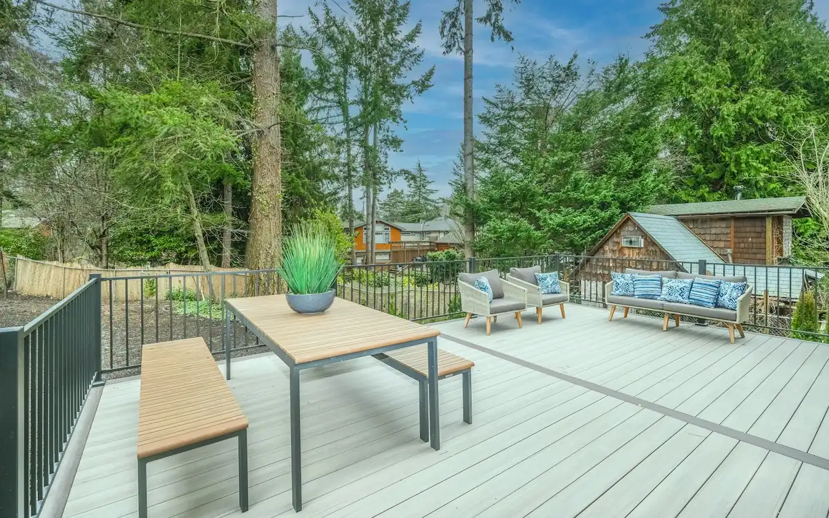A timber deck with an outdoor table, benches and cushioned seating, with a garden shed among trees behind