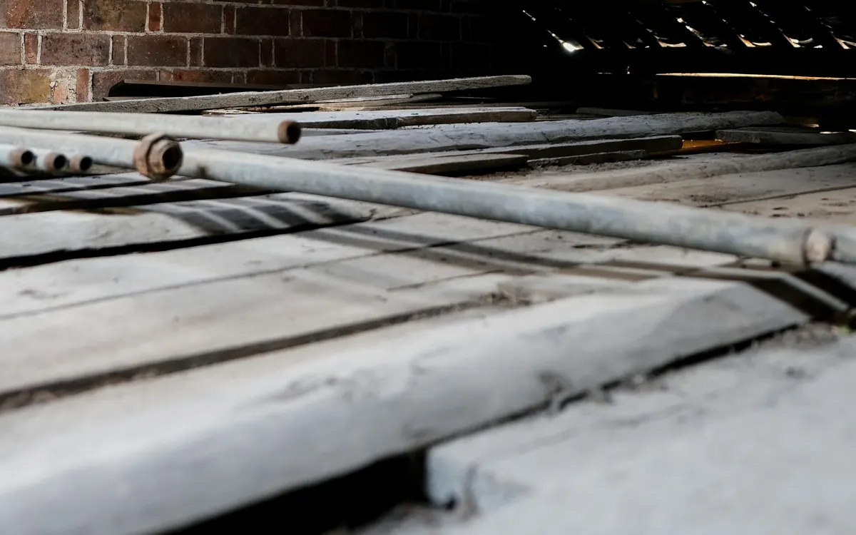 An unboarded loft floor with exposed joists, loose boards and old pipework